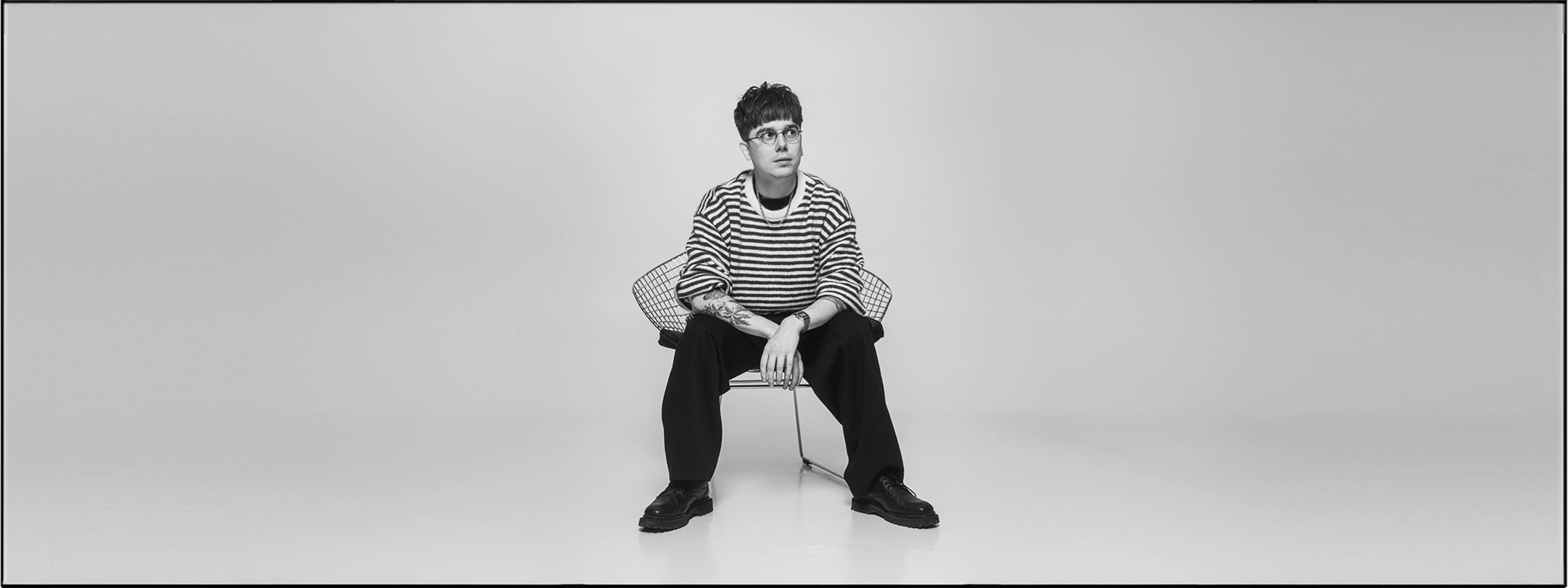 Mężczyzna w paskowanym swetrze i okrągłych okularach siedzący na metalowym krześle, z widocznym tatuażem na przedramieniu.