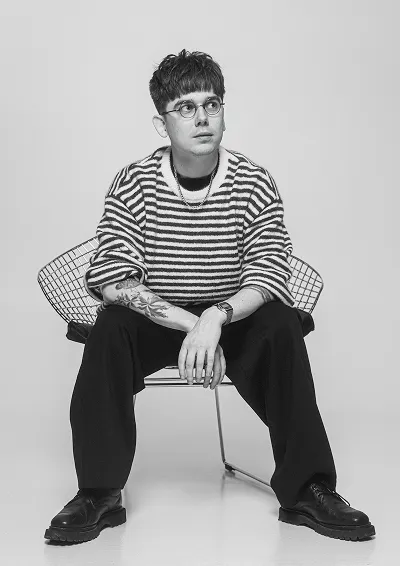 Mężczyzna w paskowanym swetrze i okrągłych okularach siedzący na metalowym krześle, z widocznym tatuażem na przedramieniu.